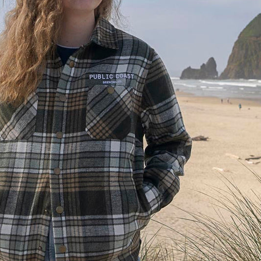 Person wearing a plaid shirt with 'Public Coast Brewing' logo on a beach